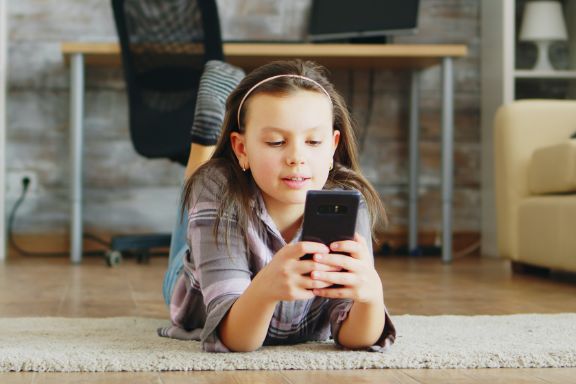 A young girl with light brown hair tied in a pink headband lies on her stomach on a beige rug in a cozy living room, happily scrolling on her smartphone. She wears a striped shirt and jeans, with a wooden desk, bookshelf, potted plant, and beige couch blurred in the background, evoking a sense of innocent curiosity in exploring social media like Bluesky.